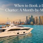 A modern, luxurious white yacht cruising on the clear blue waters of Dubai. The iconic sail-shaped Burj Al Arab hotel is visible on the left, with the Dubai city skyline in the background under a warm sunset sky. The image has a text overlay reading: 'When to Book a Dubai Yacht Charter: A Month-by-Month Guide'.